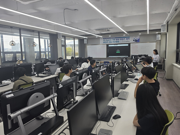 부산경상대학교 창의관 컴퓨터 실습실에서 콘텐츠 기업 재직자들이 모니터 앞에 앉아 강의를 듣고 있고, 전면 스크린에 'AI Agent 기반 영상 콘텐츠 제작 바이브 코딩 커리큘럼' 안내가 표시돼 있는 모습
